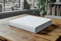 White hardcover book with blank cover on rustic wooden table in bright modern workspace with plants and natural light.