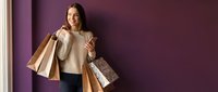 Happy young Asian woman holding multiple shopping bags after successful retail therapy session against purple wall background.