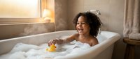 Happy African American toddler girl enjoying bubble bath with rubber duck toy in modern bathroom during golden hour sunlight streaming through window.