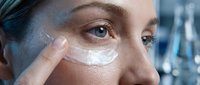 Young woman applying white moisturizing cream under eye area for skincare routine and anti-aging treatment in modern bathroom setting.
