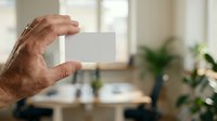 Senior male hand holding blank white card in modern office workspace with green plants and natural light, suitable for business mockups and corporate branding presentations.