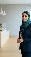 Middle Eastern woman wearing hijab and professional navy blazer smiling in modern office kitchen with pendant lighting and wooden flooring, suitable for business, corporate, and workplace diversity concepts.