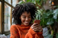 Young African American woman with natural curly hair wearing orange sweater smiling while using smartphone near window, surrounded by green plants, lifestyle portrait.