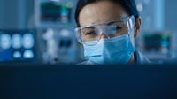 Female scientist in protective mask and safety glasses working on computer in modern laboratory conducting research and data analysis.