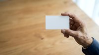 Cropped hand of mature man holding blank white business card against wooden floor background for corporate branding and identity mockup purposes.
