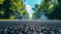 Dramatic low angle view of wet asphalt road surface with water drops after rain, blurred green forest background and rising steam creating atmospheric scene.
