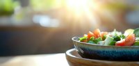 Fresh vegetable salad with tomatoes and greens in ceramic bowl on wooden table with sunlight streaming through window.