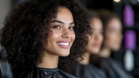 Young African American woman with curly hair smiling at hair salon, with blurred customers in background. Professional beauty service environment.