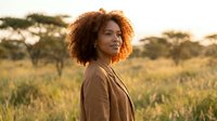 Young African woman with curly hair in beige blazer standing in golden savanna landscape at sunset, professional portrait for business and lifestyle content.