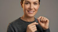 Young woman with brown hair and fair complexion wearing gray sweater, smiling while making small gesture with fingers against neutral background for advertising and lifestyle content.