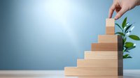 Female hand placing wooden block on top of ascending staircase structure against blue background with green plant, representing growth and business success.