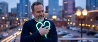Mature Caucasian businessman in suit holding glowing green heart symbol against urban city skyline at dusk, representing corporate social responsibility and care.