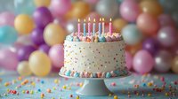 White birthday cake decorated with colorful sprinkles and lit candles on cake stand against blurred pastel balloon background with scattered confetti on surface.