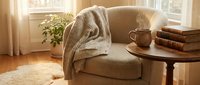 Cozy beige armchair with knitted throw blanket beside wooden side table with coffee cup and books in warm sunlit living room for home decor and relaxation.