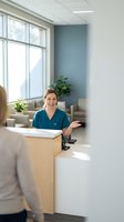 Friendly female healthcare worker in teal scrubs smiling at modern medical office reception desk with natural lighting and contemporary interior design.