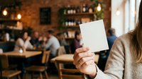 Woman holding blank white card in casual cafe setting with blurred customers and brick wall background for mockup and branding purposes.