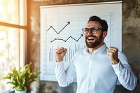 Excited Middle Eastern businessman celebrating success near whiteboard with growth charts. Positive male professional showing victory gesture in modern office.