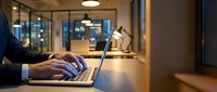 Professional businessman working late on laptop in modern office environment with warm lighting and glass partitions for corporate productivity concepts.