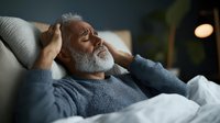 Senior African American man with gray beard experiencing headache pain while resting on couch at home, wearing casual sweater, emotional health concept.