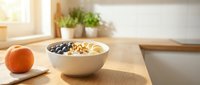 Healthy breakfast bowl with granola, blueberries, and banana slices on wooden table near window with fresh fruit and plants.