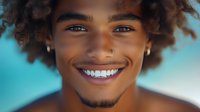 Young African American man with curly hair and bright smile showing perfect white teeth against blurred tropical beach background for summer lifestyle content.