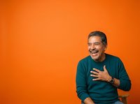 Middle-aged man with dark hair and facial stubble, appearing to be of Latin or Mediterranean descent, wearing a teal long-sleeve shirt against a vibrant orange background. He displays a genuine, warm smile while gesturing expressively with his hands, conveying enthusiasm and positivity. Suitable for corporate communications, lifestyle marketing, and motivational content.