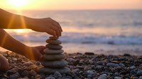 Hands carefully stacking smooth stones on pebble beach at golden sunset with ocean waves in background creating zen balance meditation scene.