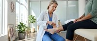 Female Caucasian physiotherapist in white coat examining patient leg in modern bright medical clinic office with plants and natural lighting.