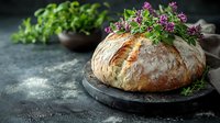 Fresh crusty artisan sourdough bread loaf garnished with purple wildflowers and herbs on dark stone board against moody rustic background with flour dust.