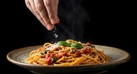 Hand sprinkling cheese over spaghetti pasta with tomato sauce and fresh basil on dark ceramic plate against black background for culinary presentation.