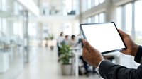 Businessman with dark skin holding blank tablet device in modern office with colleagues and windows in background.