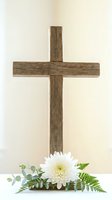 Rustic wooden cross with white chrysanthemum flower and green fern leaves on neutral background for religious ceremonies and spiritual worship concepts.