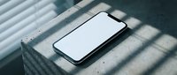 Modern smartphone with blank white screen lying on concrete surface with dramatic window shadow patterns for mockup and app presentation designs.
