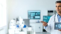 Middle-aged Caucasian male doctor in white coat holding tablet with medical scan in modern hospital laboratory with equipment and monitors in background.