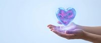 Hands holding glowing holographic heart with digital circuit patterns floating above palms against soft blue background for healthcare technology concepts.