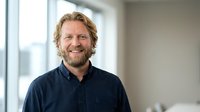 Smiling middle-aged Caucasian businessman with beard and wavy hair in navy shirt standing in modern office with natural light from windows.