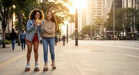 Two diverse young women friends walking together on urban city street during golden hour sunset sharing smartphone and laughing joyfully.