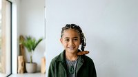 Young child with braided hair and warm smile wearing olive green jacket, standing indoors near window with potted plants, suitable for educational, family, and lifestyle content.