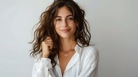 Young woman with wavy brunette hair and natural makeup wearing white shirt smiling at camera. Casual lifestyle portrait against light wall background.