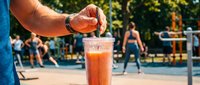 Caucasian male hand squeezing fresh lemon into refreshing beverage at outdoor summer event with blurred crowd and sports court in background.
