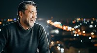 Middle-aged man with dark hair and stubble, Mediterranean ethnicity, wearing dark sweater against blurred city lights at night, looking thoughtful and contemplative for lifestyle and portrait use.