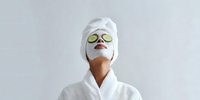 Woman in white bathrobe with facial mask and cucumber slices on eyes for spa skincare treatment isolated on gray background for wellness design.
