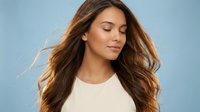 Young Hispanic woman with long flowing brown hair and closed eyes in peaceful expression wearing white top against soft blue background for wellness content.