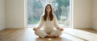 Woman with long dark hair and medium complexion in lotus position on wooden floor practicing meditation in bright minimalist room with large windows, suitable for wellness, mindfulness, yoga, and mental health content.