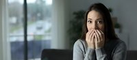 Young Asian woman covering mouth with hands showing shock or surprise emotion in modern home interior with natural window light for lifestyle concepts.