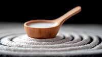 Wooden measuring spoon with white powder on circular pattern of sugar or salt, dramatic dark background with selective focus creates minimalist composition.