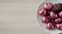 Heart shaped chocolate candies wrapped in pink and brown foil arranged on white ceramic plate against light wooden background, romantic holiday treat.