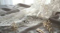 Delicate ivory lace fabric with embroidered floral details draped elegantly over soft gray silk, adorned with pearl beads and baby's breath flowers for wedding and bridal design projects.