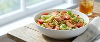 Fresh pasta salad with grilled chicken, cherry tomatoes, avocado and herbs in white bowl on wooden table near window with iced tea glass.