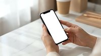 Hands holding smartphone with blank white screen mockup in cozy cafe setting with coffee cups on marble table for app presentation and digital design.